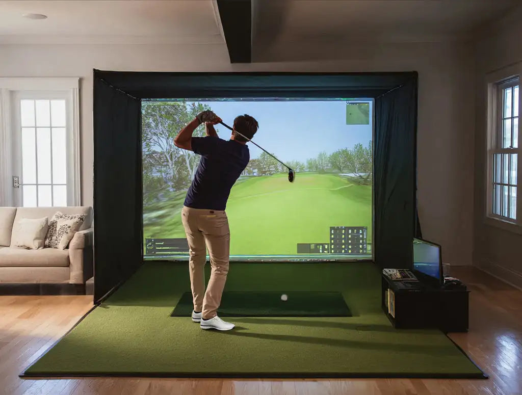 A man in a navy blue shirt and beige pants stands poised with a golf club, ready to swing at an immersive virtual green displayed on a large screen within a home indoor driving range setup.